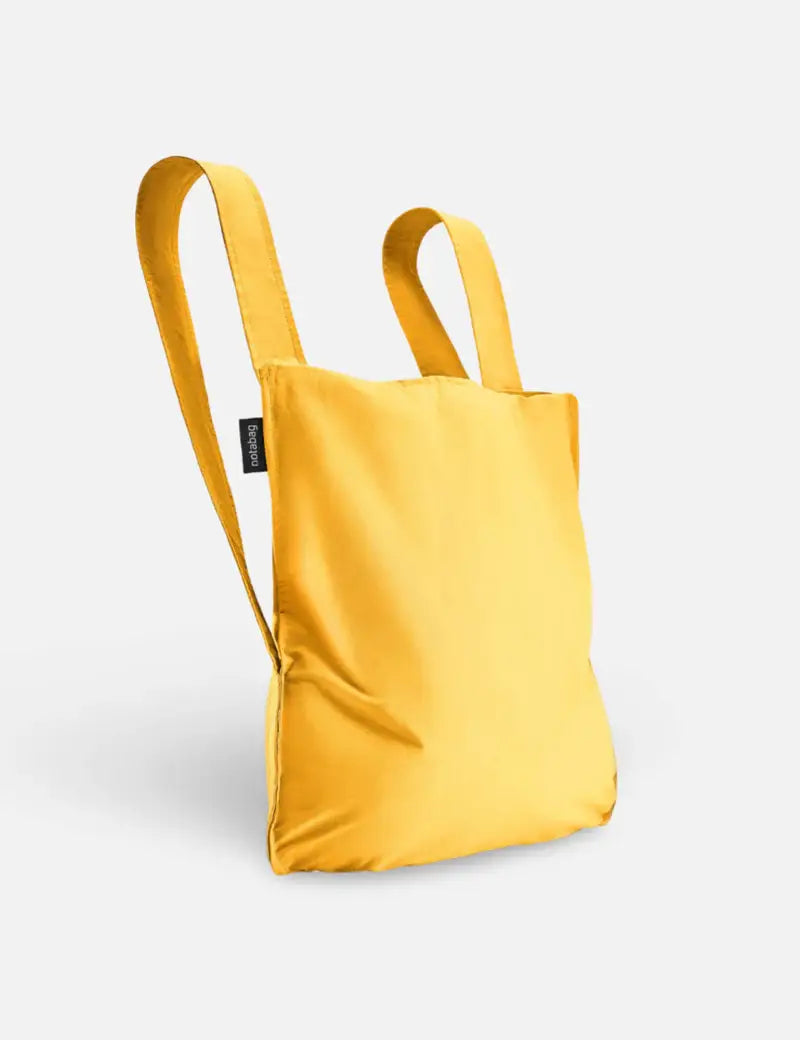 Der Notabag „Original Golden“ von Notabag ist eine leuchtend gelbe, umweltfreundliche Kombination aus Tasche und Rucksack mit zwei Schulterriemen und minimalistischem Design vor hellem Hintergrund.