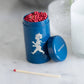 Die Solstickan Design | Streichholzdose Blau von Solstickan Design zeigt eine blaue Dose mit der weißen Silhouette eines laufenden Kindes, gefüllt mit roten Streichhölzern. Der Deckel liegt daneben, ein Streichholz liegt davor auf einer weißen Fläche.