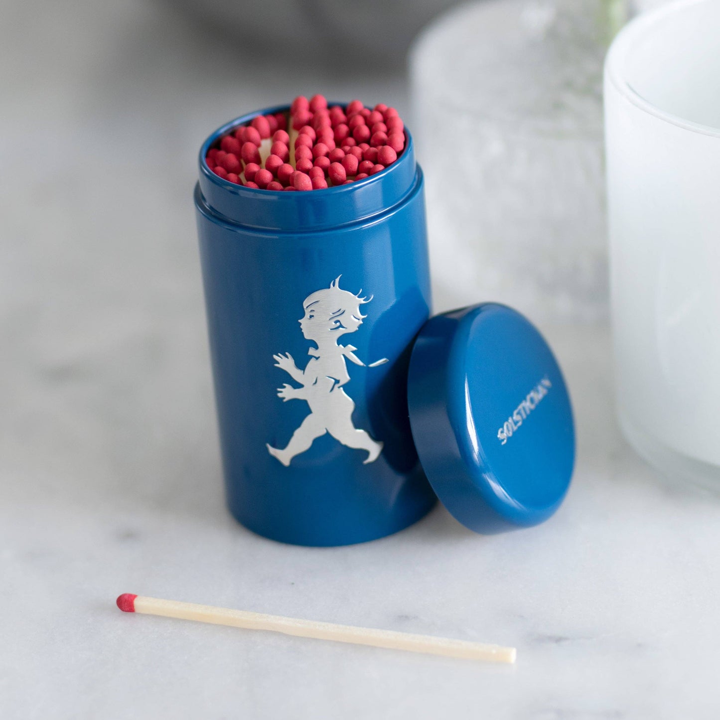 Die Solstickan Design | Streichholzdose Blau von Solstickan Design zeigt eine blaue Dose mit der weißen Silhouette eines laufenden Kindes, gefüllt mit roten Streichhölzern. Der Deckel liegt daneben, ein Streichholz liegt davor auf einer weißen Fläche.