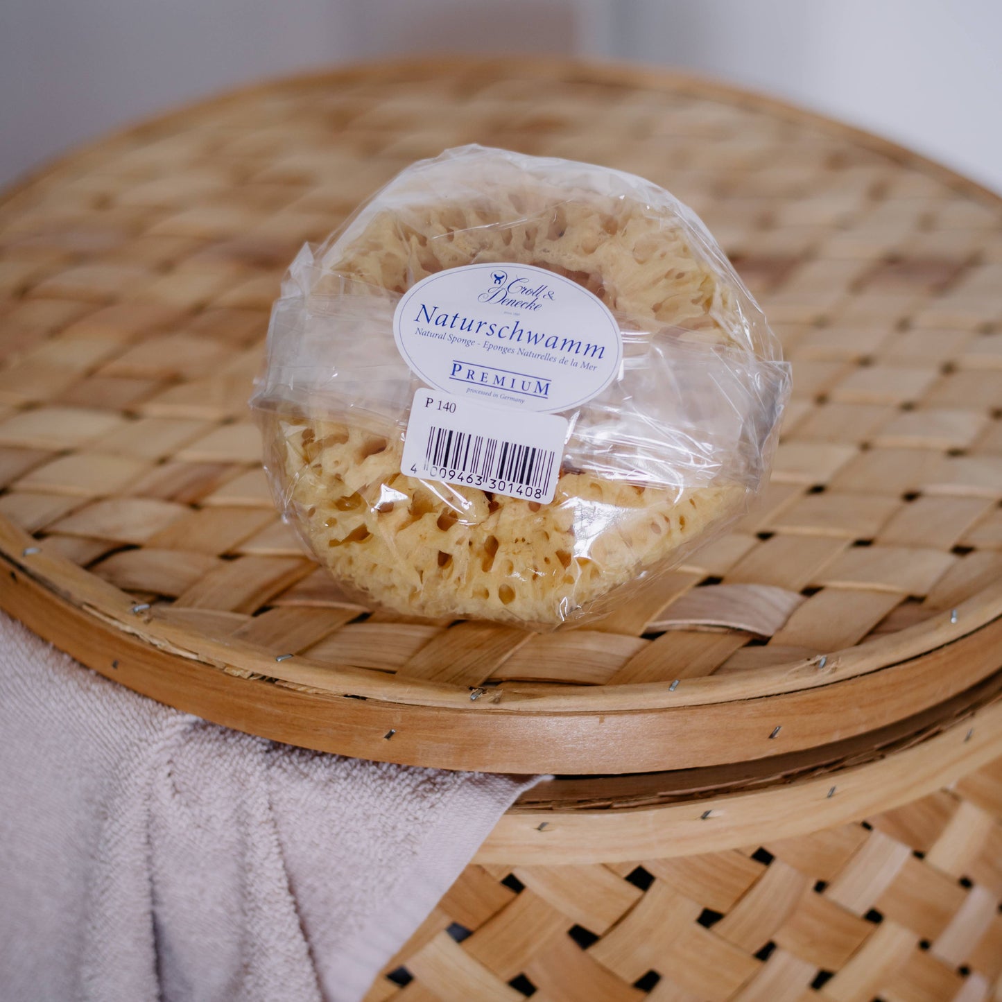 Ein Croll & Denecke Naturschwamm aus dem Meer S mit deutschem Label liegt auf einem Korb, darüber ein helles Handtuch - ideal für nachhaltige Pflege und ein luxuriöses Duscherlebnis.
