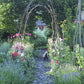 Ein Steinweg führt unter einem rustikalen Astbogen hindurch in einen blühenden Cottage-Garten – Schauplatz unseres Halbtageskurses am Samstag, 27.2.2027 | 14.00 – 17.00 Uhr: Der Cottage-Garten: Der Garten der Zukunft.