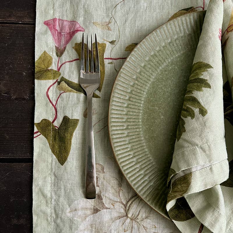 Auf dem Leinen-Tischläufer „Flora Danica Atlas“ von Koustrupco (145 x 45 cm) liegen eine grüne Serviette mit Blumenmuster und ein Teller, links davon eine silberne Gabel – so entsteht auf der dunklen Holzoberfläche eine bezaubernde Tischdekoration.