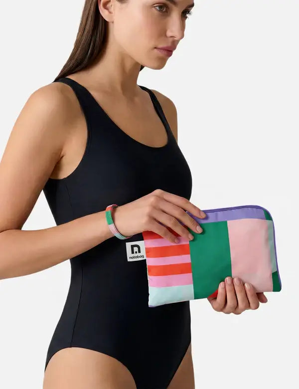 Eine Frau in einem schwarzen einteiligen Badeanzug hält das Notabag 'Flow' Handtuch 78 x 160 cm, ein schnell trocknendes Recycling-Handtuch von Notabag mit einem geometrischen Streifenmuster in Grün, Rosa, Lila und Rot. Dazu trägt sie ein passendes Armband.