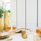 Ein modernes Esstisch-Set mit der goldenen Vase AULICA | Glänzende, goldenem Geschirr, erröteten Gläsern, cremefarbenen Servietten und grünem Laub auf einem weißen Marmortisch für einen eleganten und luxuriösen Look.