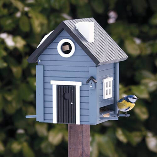 Ein stilvoller blauer „Nistkasten & Futterhaus“ von Wildlife Garden in Form eines Hauses steht auf einem Pfosten, an dessen Seite ein bezaubernder blau-gelber Vogel sitzt. Grüne Blätter füllen den sanft unscharfen Hintergrund aus.