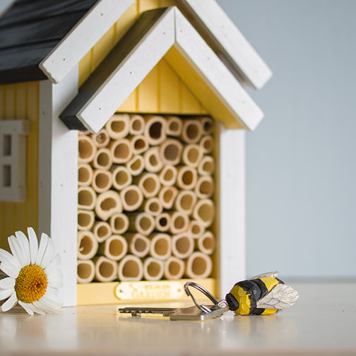 Ein gelb-weißes Bienenhaus mit Bambusrohren steht auf einem Tisch neben einer Gänseblümchenblüte und dem handgefertigten Schlüsselanhänger aus Holz 'Hummel' von Wildlife Garden.