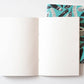Ein aufgeschlagenes Notizbuch ruht auf einem geschlossenen Papaya Paper Products Notizheft A5 mit Federmuster blau, das ein schwarz-weißes Federmuster auf einem blaugrünen Einband zeigt. Beide sind aus Munken Print Cream Papier auf weißem Hintergrund hergestellt.