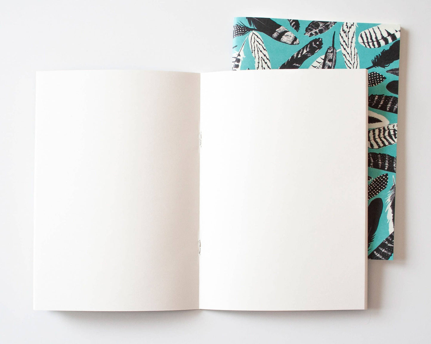 Ein aufgeschlagenes Notizbuch ruht auf einem geschlossenen Papaya Paper Products Notizheft A5 mit Federmuster blau, das ein schwarz-weißes Federmuster auf einem blaugrünen Einband zeigt. Beide sind aus Munken Print Cream Papier auf weißem Hintergrund hergestellt.