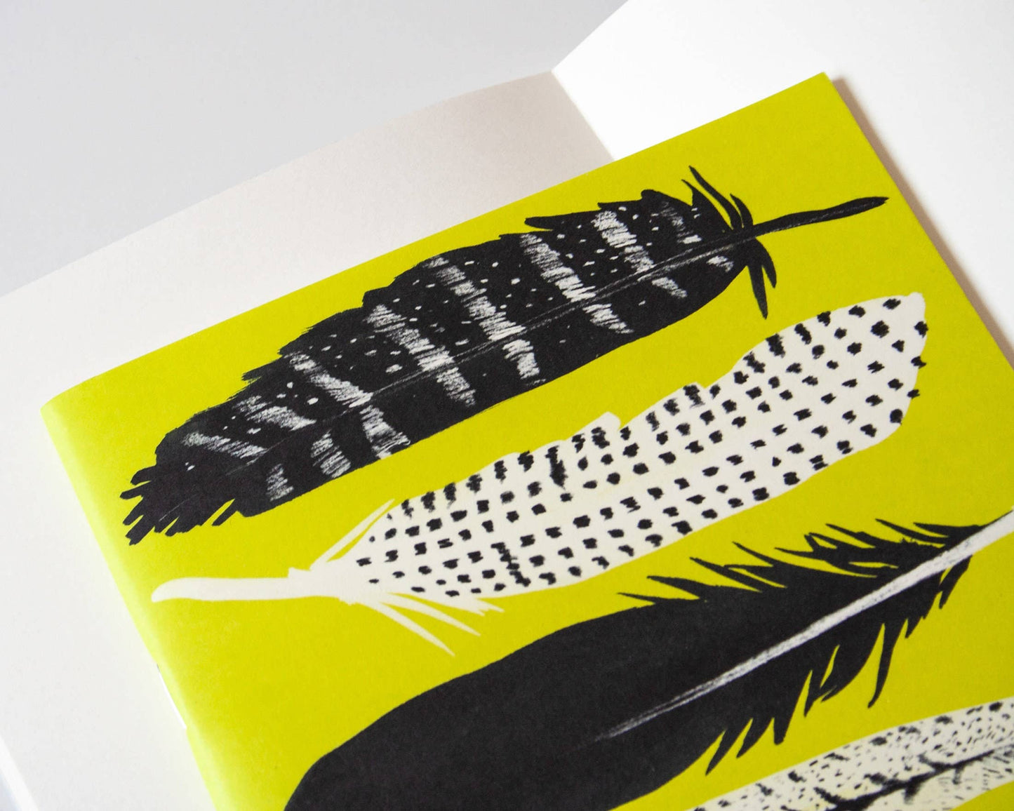 Eine Nahaufnahme des Papaya Paper Products | Schönes Notizheft A5 mit Federn in gelb zeigt schwarz-weiß illustrierte Federn mit verschiedenen Mustern auf einem leuchtend gelben Einband, hergestellt von Papaya Paper Products.