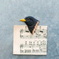 Mit dem Wildlife Garden | 'Amsel' Magnet aus handgeschnitztem Holz wird ein Notenblatt auf eine graue Fläche gepinnt, so dass die obere linke Ecke wie der Körper des Vogels mit seinem gelben Schnabel und den schwarzen Federn aussieht.