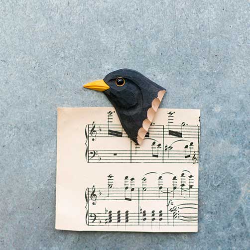 Mit dem Wildlife Garden | 'Amsel' Magnet aus handgeschnitztem Holz wird ein Notenblatt auf eine graue Fläche gepinnt, so dass die obere linke Ecke wie der Körper des Vogels mit seinem gelben Schnabel und den schwarzen Federn aussieht.