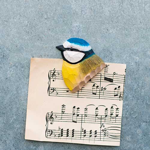 A Wildlife Garden 'Blaumeise' Magnet aus handgeschnitztem Holz in Gelb und Blau liegt auf einem Notenblatt auf einer strukturierten grauen Fläche.