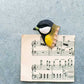 A Wildlife Garden 'Kohlmeise' Magnet aus handgeschnitztem Holz, in gelb, schwarz und weiß, hält ein Notenblatt vor grauem Hintergrund. Der Clip ist vogelförmig und von Gartenvögeln inspiriert.