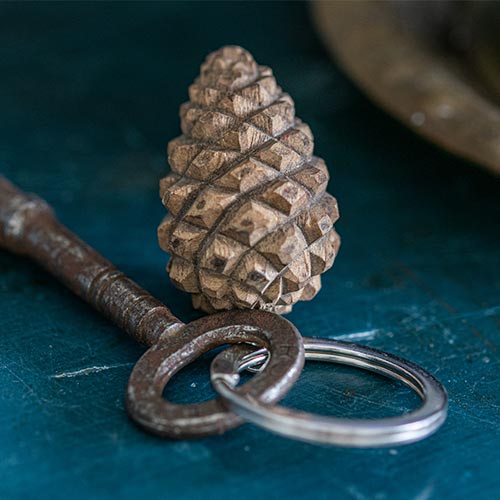 Eine Nahaufnahme eines Metallschlüssels an einem Schlüsselanhänger mit dem Wildlife Garden handgefertigter Schlüsselanhänger aus Holz, Zapfen - ein kunstvoll geschnitzter Kiefernzapfen - auf einer dunkelblauen Oberfläche. Perfekt als Geschenk für Naturfreunde.