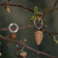 Der Wildlife Garden Handgefertigter Schlüsselanhänger aus Holz "Zapfen" von Wildlife Garden hängt an einem kleinen Zweig mit grünen Blättern - ein ideales Geschenk für Naturfreunde. Im Hintergrund sieht man weitere Anhänger und Äste.