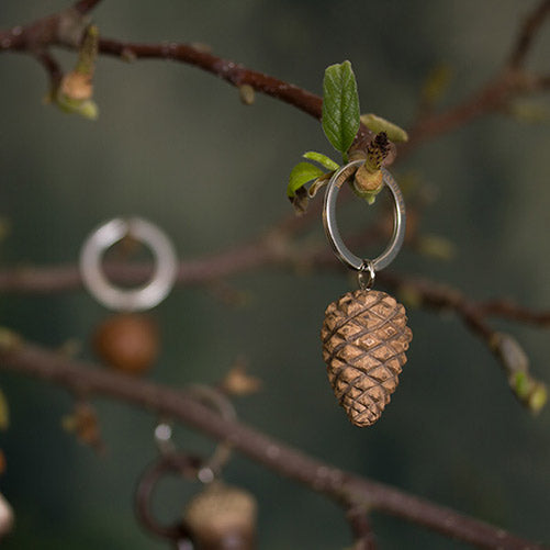 Der Wildlife Garden Handgefertigter Schlüsselanhänger aus Holz "Zapfen" von Wildlife Garden hängt an einem kleinen Zweig mit grünen Blättern - ein ideales Geschenk für Naturfreunde. Im Hintergrund sieht man weitere Anhänger und Äste.