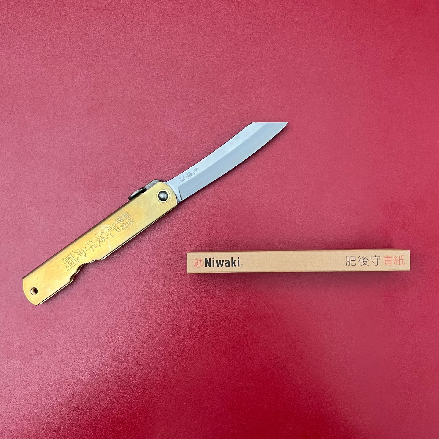 Ein Niwaki | 'Blue Steel Higonokami' Klappmesser mit einem in japanische Schriftzeichen eingravierten Messinggriff liegt geöffnet auf einer roten Fläche neben einer rechteckigen Pappschachtel mit der Aufschrift Niwaki.