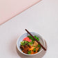 Eine lebendige Ramen-Schale mit Nudeln, geschnittenem Fleisch, eingelegtem Ingwer, Erbsen, Kräutern und Chiliflocken wird mit White Mausu | Erdnuss Rayu Sauce 440 g von White Mausu Ltd übergossen und mit Stäbchen auf einer Matte über einer hellen Arbeitsfläche serviert.