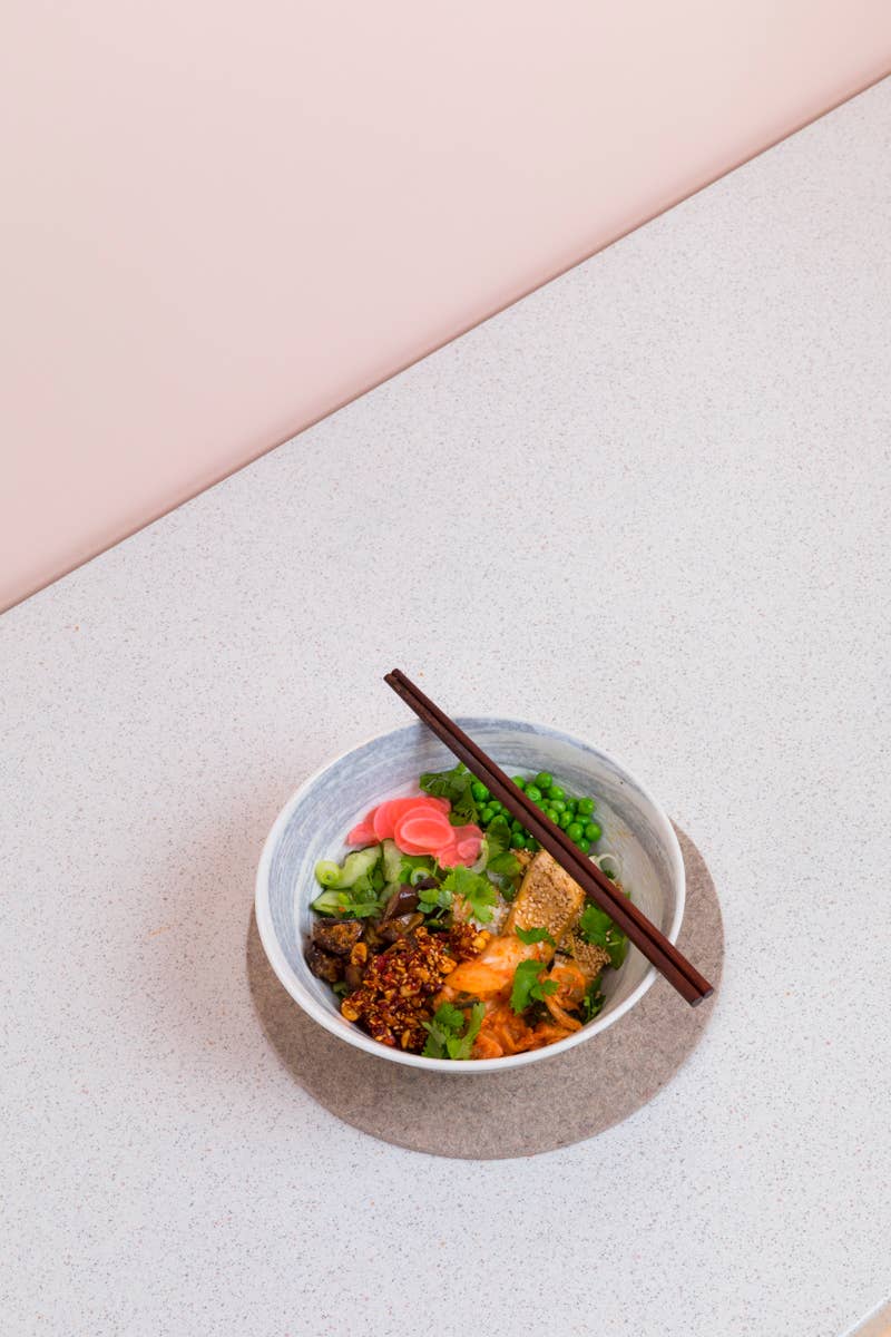 Eine lebendige Ramen-Schale mit Nudeln, geschnittenem Fleisch, eingelegtem Ingwer, Erbsen, Kräutern und Chiliflocken wird mit White Mausu | Erdnuss Rayu Sauce 440 g von White Mausu Ltd übergossen und mit Stäbchen auf einer Matte über einer hellen Arbeitsfläche serviert.