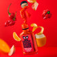 Eine Flasche Crazy Bastard Sauce Trinidad Scorpion & Clementine 100 ml scharfe Soße hebt sich von einem leuchtend roten Hintergrund ab, umgeben von schwimmenden Trinidad Scorpion Chilischoten, Apfelscheiben und Clementinenschalen.