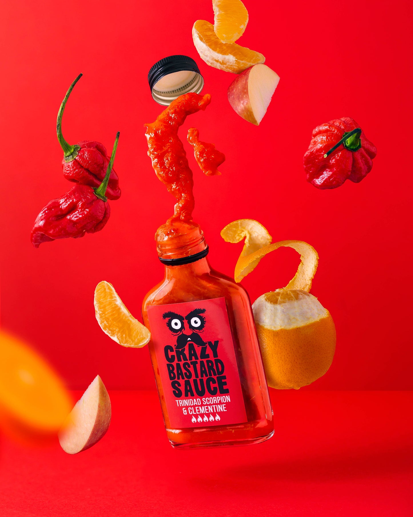 Eine Flasche Crazy Bastard Sauce Trinidad Scorpion & Clementine 100 ml scharfe Soße hebt sich von einem leuchtend roten Hintergrund ab, umgeben von schwimmenden Trinidad Scorpion Chilischoten, Apfelscheiben und Clementinenschalen.