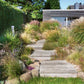 Ein Weg aus breiten hölzernen Trittsteinen schlängelt sich durch Gräser und Blumen und führt zu einem modernen Holzhaus - die perfekte Inspiration für Ihren Traumgarten aus "Die schönsten Terrassen und Sitzplätze" vom Callwey Verlag.