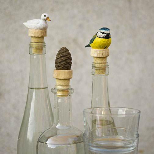 Drei Glasflaschen mit handgefertigten Korkverschlüssen - eine mit einer weißen Ente, eine mit dem handgeschnitzten Holzflaschenverschluss 'Tannenzapfen' von Wildlife Garden und eine mit einem blau-gelben Vogel, davor ein Klarglasbecher.