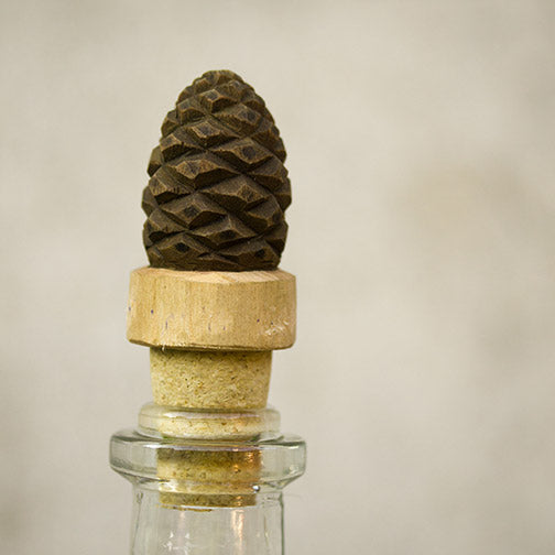 Eine Glasflasche wird mit dem Wildlife Garden Handgeschnitzter Flaschenverschluss aus Holz 'Tannenzapfen' verschlossen, der ein dekoratives, dunkelbraunes Ornament in Form eines Tannenzapfens auf einem Holzsockel vor einem schlichten, neutralen Hintergrund aufweist.