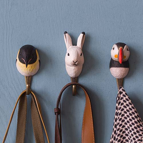Drei Wandhaken - ein Vogel, ein Wildlife Garden Handgeschnitzter Wandhaken aus Holz 'Schneehase' und ein Papageientaucher - halten Gegenstände an einer blauen Wand: Der Vogel hält einen Gurt, der Schneehase einen braunen Gurt und der Papageientaucher ein kariertes Tuch.