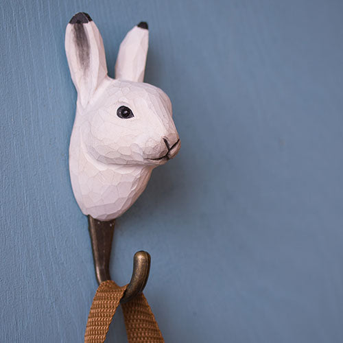 Der Wildlife Garden | Handgeschnitzter Wandhaken aus Holz 'Schneehase' von Wildlife Garden ist an einer blauen Wand montiert. Der weiße Hasenkopf mit Metallhaken eignet sich ideal als originelles Geschenk für jeden Wohnraum.