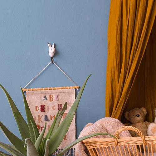 Schaffen Sie eine Kuschelecke mit blauer Wand, senfgelbem Vorhang, Weidenkorb mit Plüschbär und Decke, Aloe-Pflanze und einem Alphabet-Wandbehang auf dem handgeschnitzten Wandhaken aus Holz 'Schneehase' von Wildlife Garden - eine einzigartige Geschenkidee.
