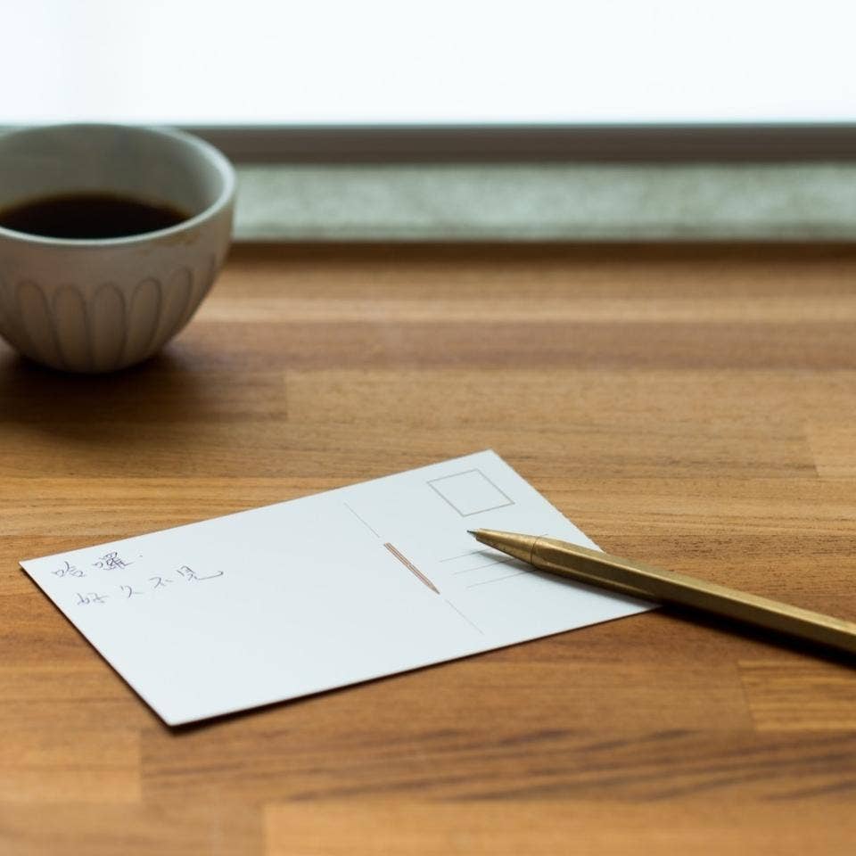Eine leere Postkarte mit handgeschriebenem japanischem Text liegt auf einem Holztisch neben einer ENTREPÔT YSTUDIO Klassischer Revolve Kugelschreiber Feder (Messing); eine Tasse Kaffee steht daneben, und durch ein Fenster im Hintergrund fällt sanftes Tageslicht.