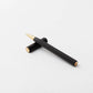 Der YSTUDIO | Harz-Rollerball-Stift (Schwarz) von ENTREPÔT, ein schwarzer, sechseckiger Stift mit goldenen Akzenten aus Acryl und Messing, ruht diagonal auf einer weißen Fläche, die Kappe liegt waagerecht unter ihm.