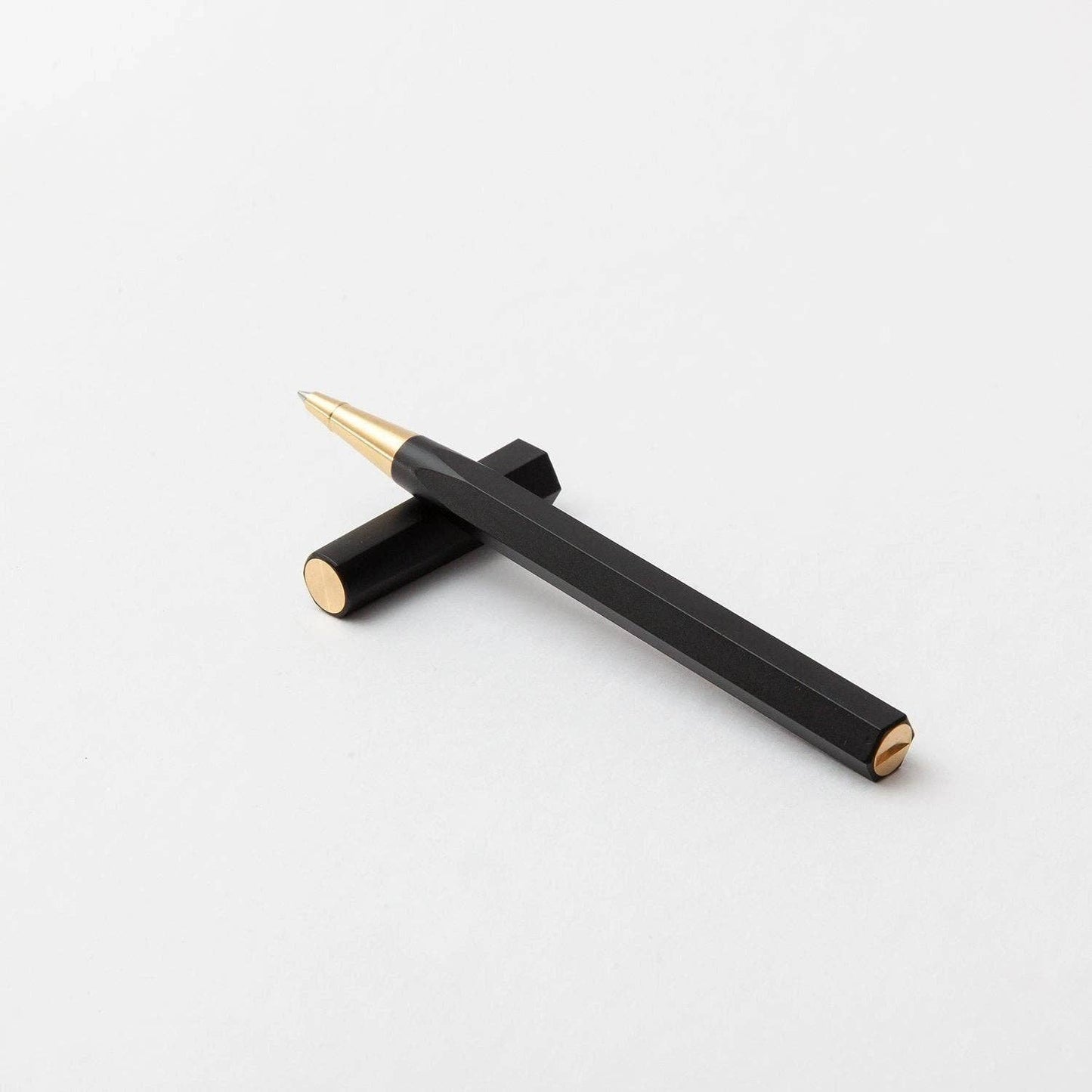 Der YSTUDIO | Harz-Rollerball-Stift (Schwarz) von ENTREPÔT, ein schwarzer, sechseckiger Stift mit goldenen Akzenten aus Acryl und Messing, ruht diagonal auf einer weißen Fläche, die Kappe liegt waagerecht unter ihm.