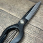 Eine Nahaufnahme der Niwaki 'Sentei Topiary Clippers' Buchsbaumschere mit glattem schwarzem Metall und glänzender Klinge mit japanischer Gravur, die auf einer hölzernen Oberfläche ruht, wobei der Griff teilweise sichtbar ist.