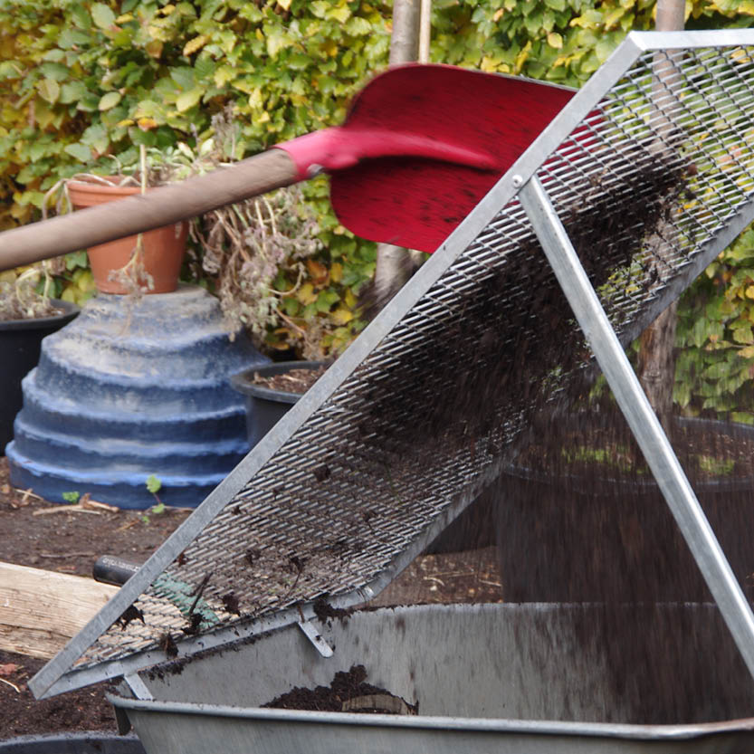 Eine rote Schaufel wirft Erde auf ein Metallgitter über einer Schubkarre während des Kurses Di., 2.3.2027 | 15.00 – 16.30 Uhr RUNDGANG: Was im März im Garten zu tun ist der Königlichen Gartenakademie.