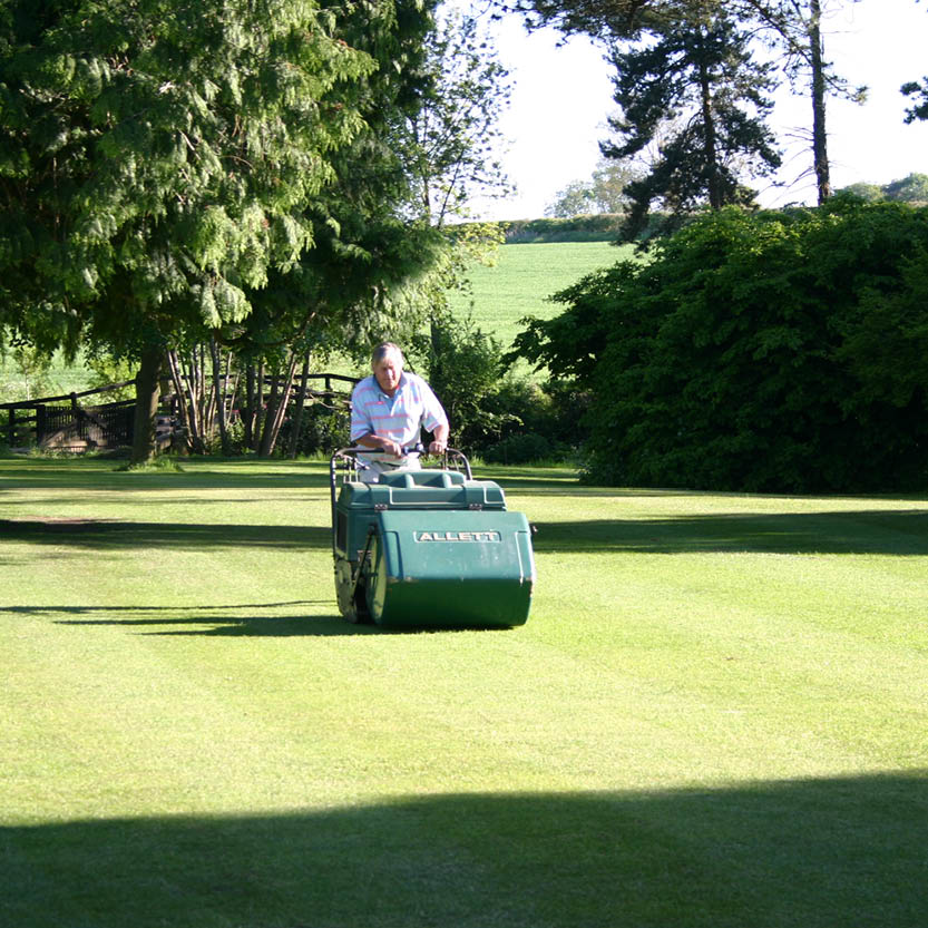 Eine Person mäht mit einem Rasenmäher das Gras in einem sonnenbeschienenen, von Bäumen umgebenen Garten - eine ideale Szene für die Veranstaltung der Königlichen Gartenakademie am Mi. 01.07.2026 | 15.00-16.30 RUNDGANG: Was zu tun ist im Garten im Juli Veranstaltung.