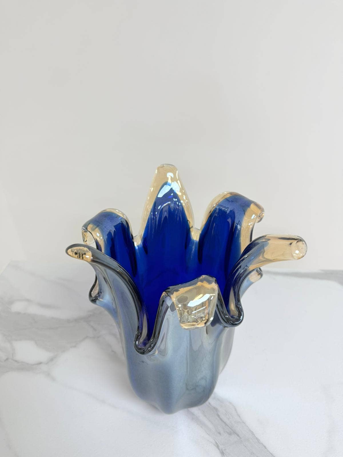 Eine blaue „Giusy“-Vase der Serie „Murano Dream“ (H 24, Ø 19 cm) aus glänzendem, spiegelndem Muranoglas mit gewellten Rändern steht auf einer weißen Marmorplatte und erinnert an eine blühende Blume.