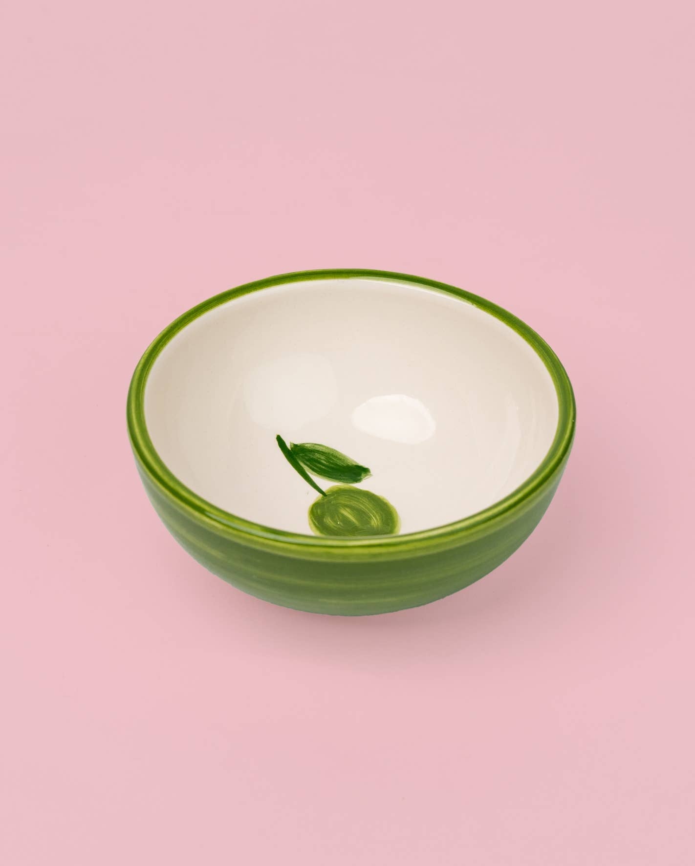 Die kleine Keramikschale „Olive“ von Alfar Tierra Cocida präsentiert sich in Grün und Weiß mit einem filigranen Blatt- und Fruchtmotiv auf der Innenseite, handgefertigt in Spanien und auf einem zarten rosafarbenen Hintergrund.