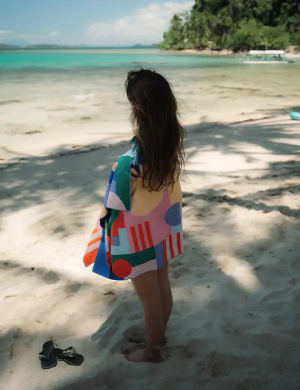 Ein Kind mit langen Haaren steht barfuß an einem Sandstrand, eingewickelt in ein Notabag | 'Flow' Handtuch 78 x 160 cm von Notabag, und blickt auf das türkisfarbene Meer. In der Nähe befinden sich Sandalen, tropische Bäume und ein Boot im Hintergrund.