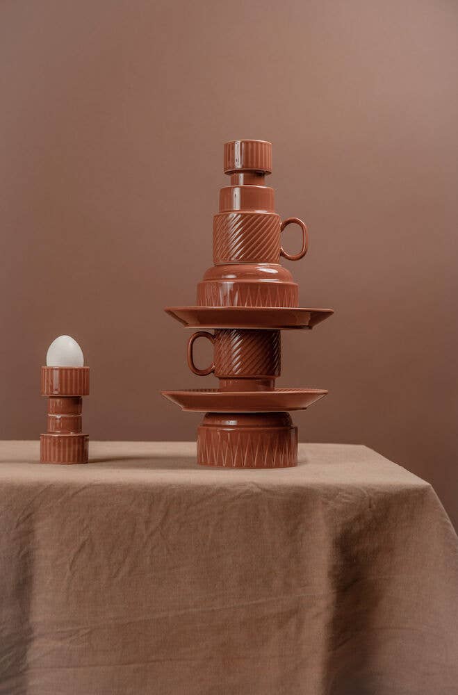 Ein Paar Kaffeebecher der Sagaform GmbH aus Terrakotta steht auf einem braunen, stoffbezogenen Tisch, daneben ein passender Eierbecher, alles vor einem einfarbig braunen Hintergrund.