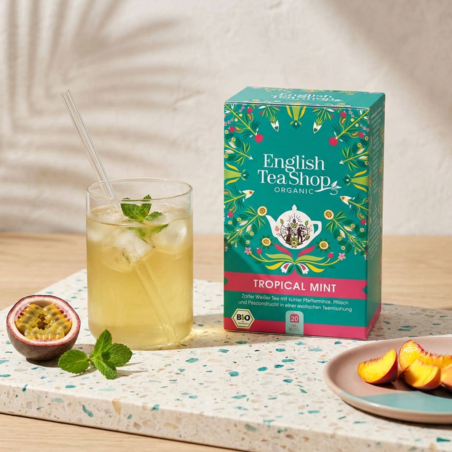 Ein Glas Eistee mit Minze steht neben einer Schachtel der Royal Nature GmbH & Co. KG English Tea Shop | Tropical Mint, BIO, 20 Teebeutel, inmitten von frischen Pfirsichscheiben und Maracuja auf einer Terrazzofläche.