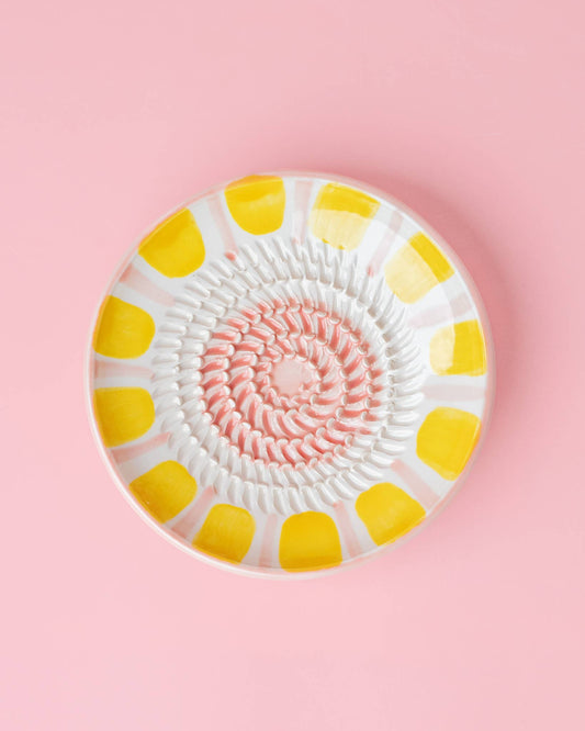 Die Alfar Tierra Cocida | Kleine Keramikreibeplatte Pastellrosa VALENTINA ist eine handgefertigte Rundreibe im Tellerstil mit pink-weißer Spirale und gelben Ovalen - ein charmantes Küchenutensil, tolles Geschenk und auf solidem Pink präsentiert.