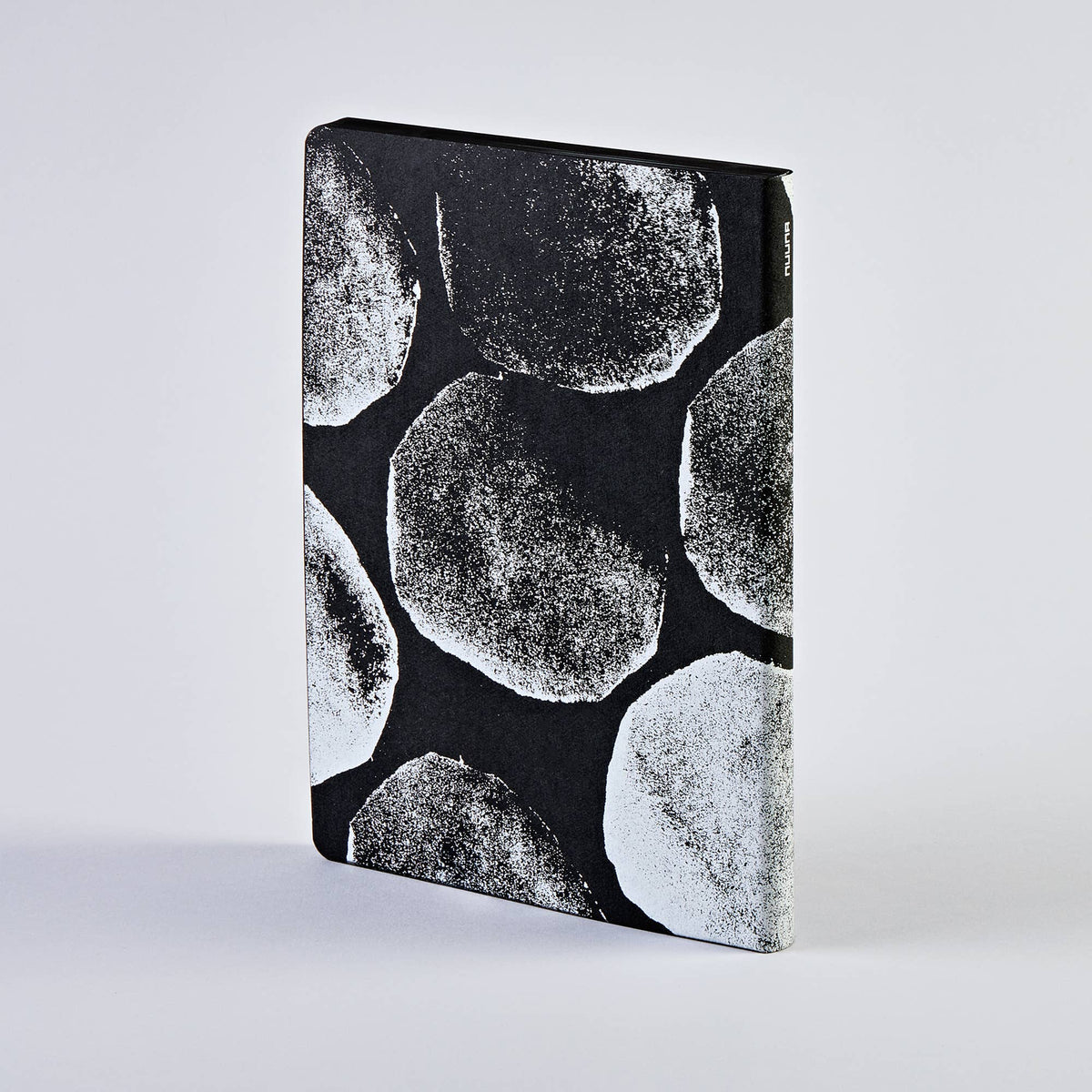 Ein schwarzes nuuna „Genesis“-Notizbuch „Not Ordinary L Light“ im Format A5+ mit weißem Kreismuster, das aufrecht vor einem schlichten hellgrauen Hintergrund steht. Dieses hochwertige Notizbuch von nuuna verfügt über charakteristische Seiten mit gepunktetem Raster und eignet sich somit perfekt für stilvolle und übersichtliche Notizen.