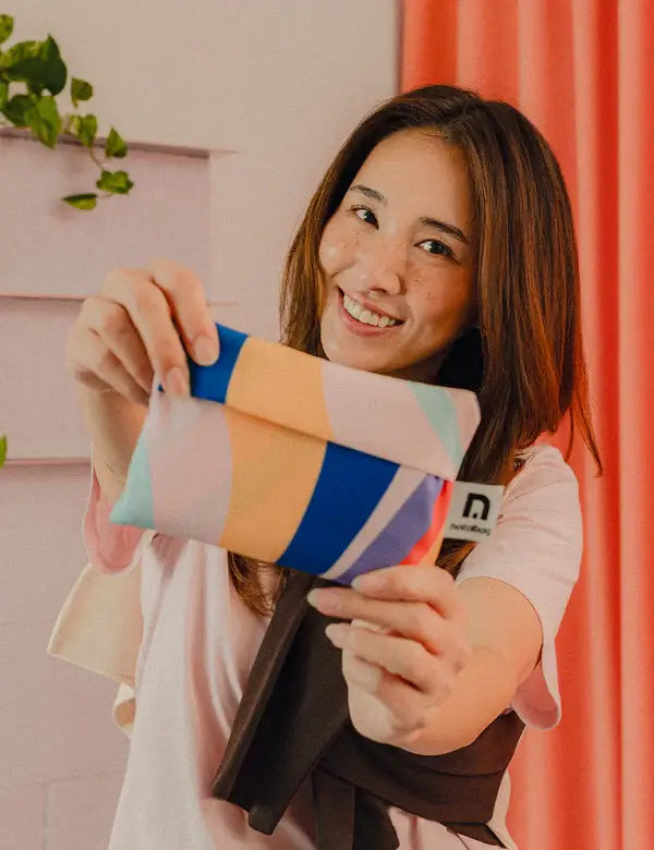 Eine lächelnde Frau mit langen braunen Haaren hält die Notabag | Shopping Tote 'Flow' von Notabag in die Kamera. Sie steht in einem Haus in der Nähe eines rosafarbenen Vorhangs, der im Hintergrund mit grünen Blättern verziert ist.