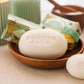 Ein weißes ovales Stück Castelbel Seife 'White Crane' Yuzu, Vetiver & Mandarine 150 g mit der Prägung "PORTUS CALE" liegt in einer Holzschale, im Hintergrund eine weitere Castelbel Seife in grünem und gelbem Papier verpackt.