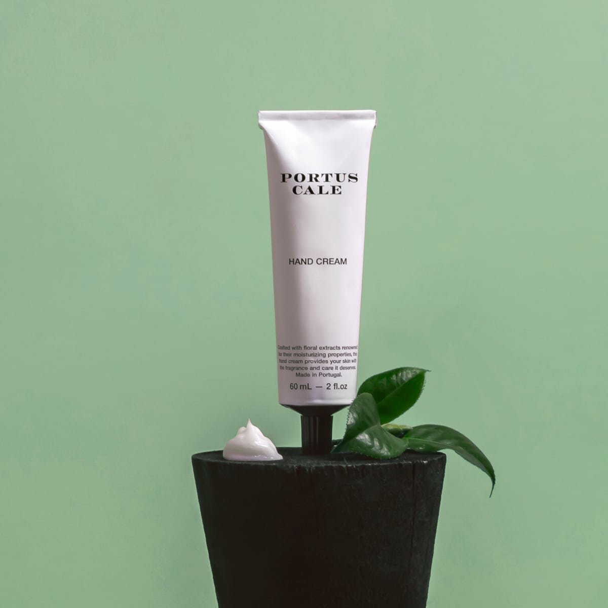 Eine weiße Tube Castelbel Handcreme White Crane Yuzu, Vetiver & Mandarinenduft 60 ml steht aufrecht auf einem schwarzen Topf mit grünen Blättern und einem Klecks Creme vor einem blassgrünen Hintergrund.