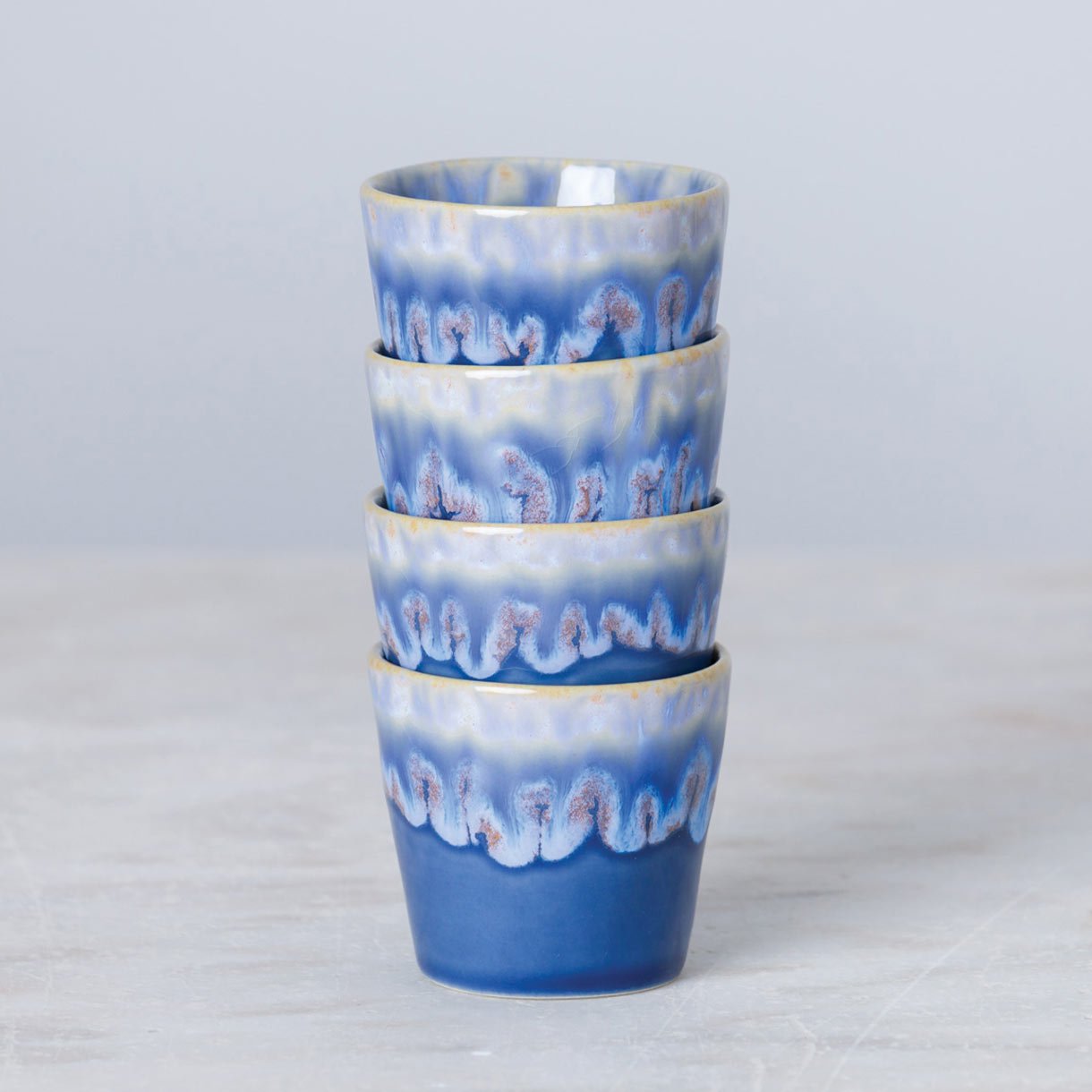 Vier Costa Nova 'Grespresso' Espressotassen Denim Blau aus Steingut und Keramik (9 cl) mit gewellter, blauer Glasur stehen auf einer hellgrauen Fläche vor hellem Hintergrund.