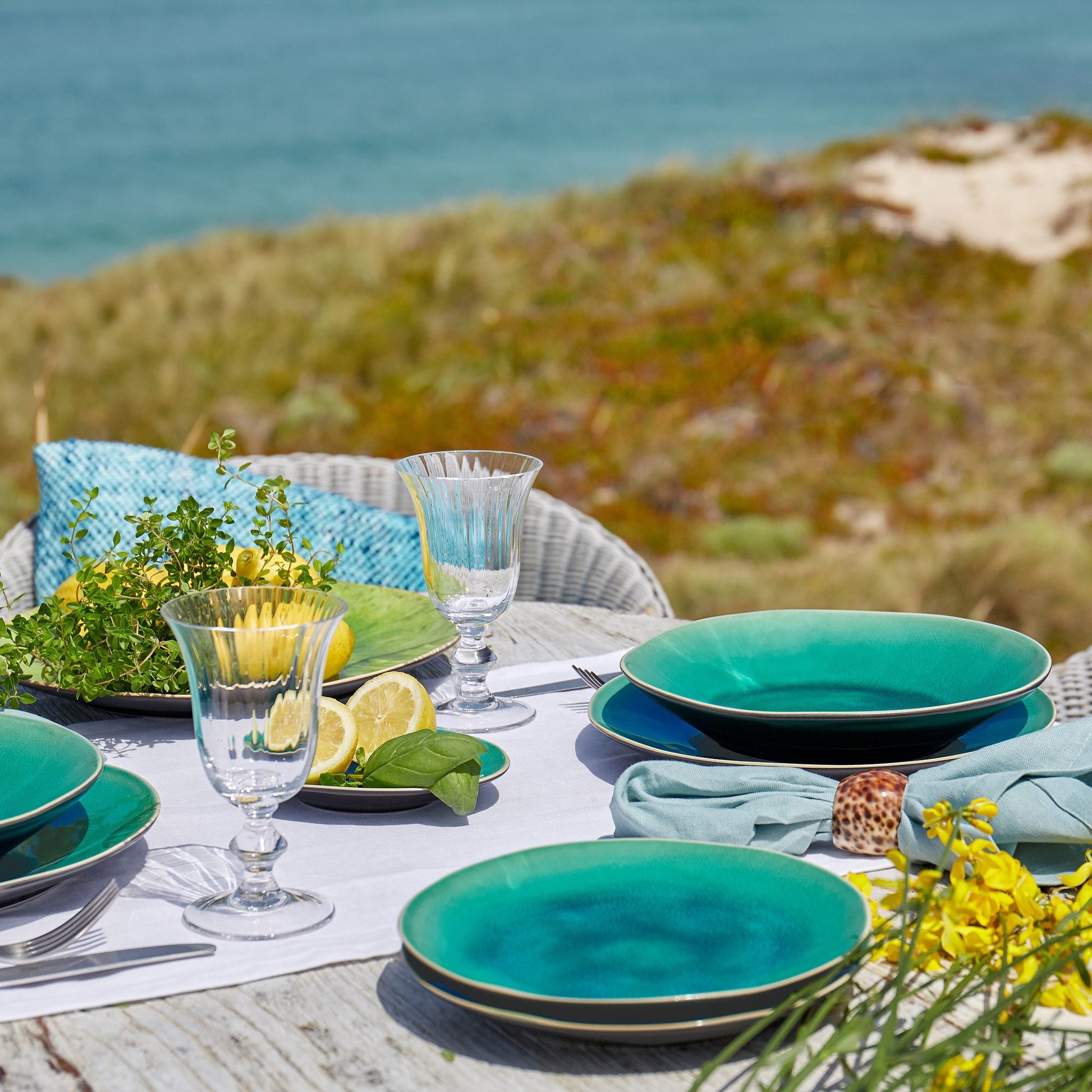 Auf einem Küstentisch stehen Costa Nova 'Azurblau' Salatteller aus Keramik 21cm, Kristallgläser, Servietten, Zitronen, Kräuter und gelbe Blumen. Im Hintergrund erscheinen Sanddünen und das Meer unter einem klaren Himmel.