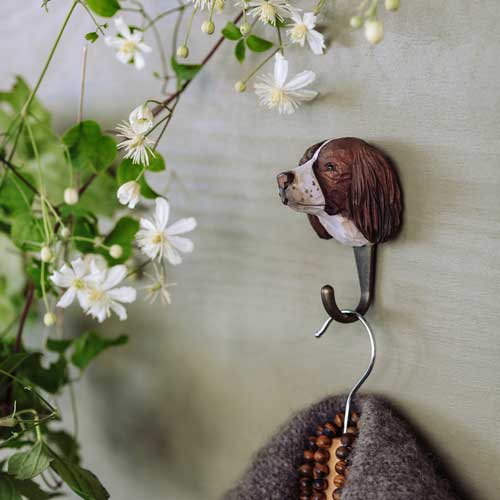 The Wildlife Garden | Handgeschnitzter Wandhaken aus Lindenholz 'Springer Spanier' zeigt einen braun-weißen Springer-Spaniel-Kopf, der einen grauen Mantel und eine Perlenkette hält. Dahinter sind weiße Blumen und grüne Blätter dekoriert.